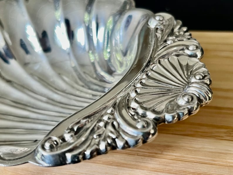 May include: A silver shell-shaped dish with a decorative, ornate rim. The dish is made of polished silver and has a reflective surface.