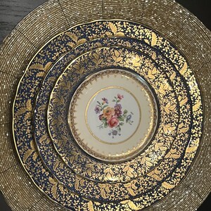 May include: A set of fine china plates arranged on a gold beaded placemat. The plates feature a navy blue and gold leaf pattern, with a smaller cream-colored plate in the center, adorned with a floral design. A sophisticated table setting.