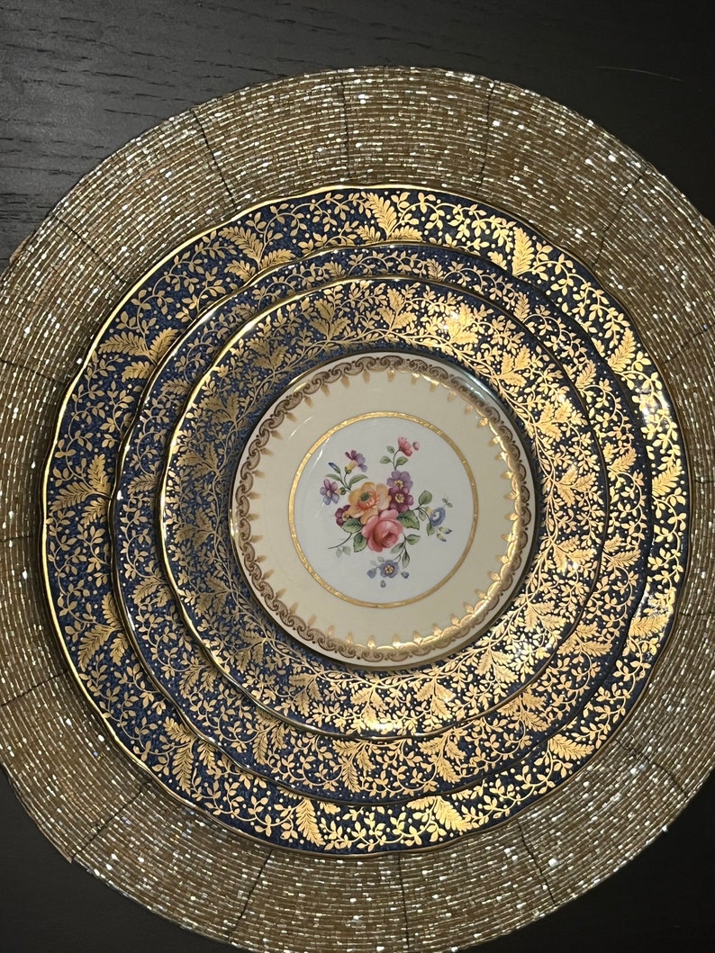 May include: A set of fine china plates arranged on a gold beaded placemat. The plates feature a navy blue and gold leaf pattern, with a smaller cream-colored plate in the center, adorned with a floral design. A sophisticated table setting.