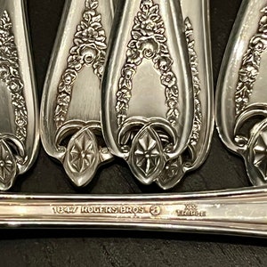 May include: A set of silver-plated spoons featuring an embossed floral garland pattern on the handles. The base of the handle is stamped with the text 1847 ROGERS BROS and XS TRIPLE. The metal has a polished finish with intricate raised detailing.