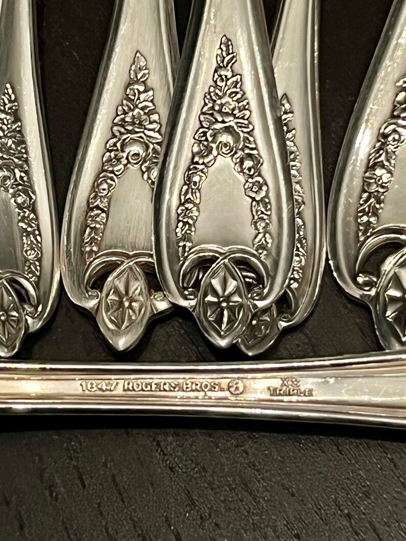 May include: A set of silver-plated spoons featuring an embossed floral garland pattern on the handles. The base of the handle is stamped with the text 1847 ROGERS BROS and XS TRIPLE. The metal has a polished finish with intricate raised detailing.