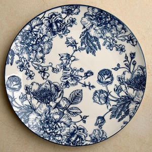 May include: A round ceramic plate featuring a cream-colored base with a detailed dark blue floral pattern. The design consists of various blooming flowers, buds, and leaves arranged across the surface, finished with a thin dark blue rim.