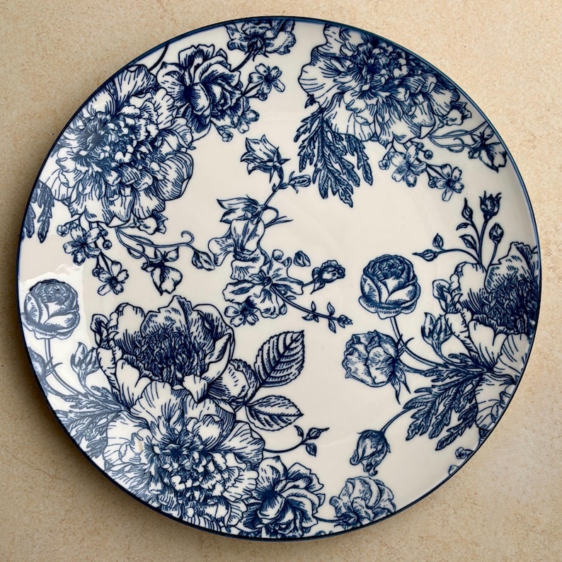 May include: A round ceramic plate featuring a cream-colored base with a detailed dark blue floral pattern. The design consists of various blooming flowers, buds, and leaves arranged across the surface, finished with a thin dark blue rim.