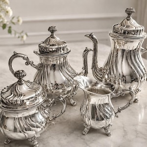 May include: A four-piece silver tea set featuring a large teapot, a smaller teapot, a lidded sugar bowl, and a creamer. Each piece has a polished, reflective finish with ornate, swirling embossed patterns, curved handles, and decorative feet.
