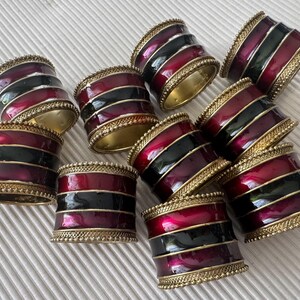 May include: A collection of decorative napkin rings. Each ring features alternating bands of burgundy and black enamel, accented with gold-coloured trim. The rings are arranged on a textured white surface.