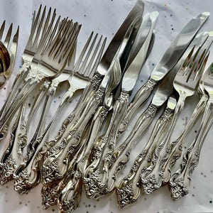 May include: A collection of ornate silver-colored flatware, including forks, knives, and a serving spoon. The silverware features detailed floral and scrollwork patterns on the handles, creating an elegant and classic aesthetic. The arrangement suggests a formal dining setting.