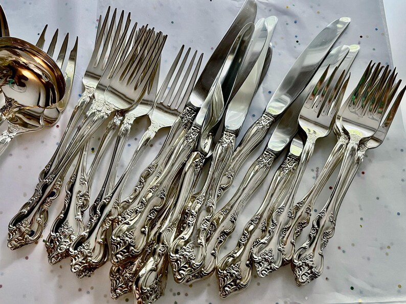 May include: A collection of ornate silver-colored flatware, including forks, knives, and a serving spoon. The silverware features detailed floral and scrollwork patterns on the handles, creating an elegant and classic aesthetic. The arrangement suggests a formal dining setting.