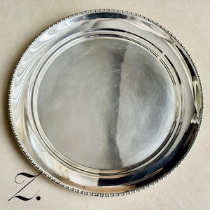 May include: A circular silver-toned metal serving tray with a polished, reflective surface and a decorative beaded edge detail. The tray features a smooth, flat center area surrounded by a slightly raised rim with a textured, repeating bead pattern.