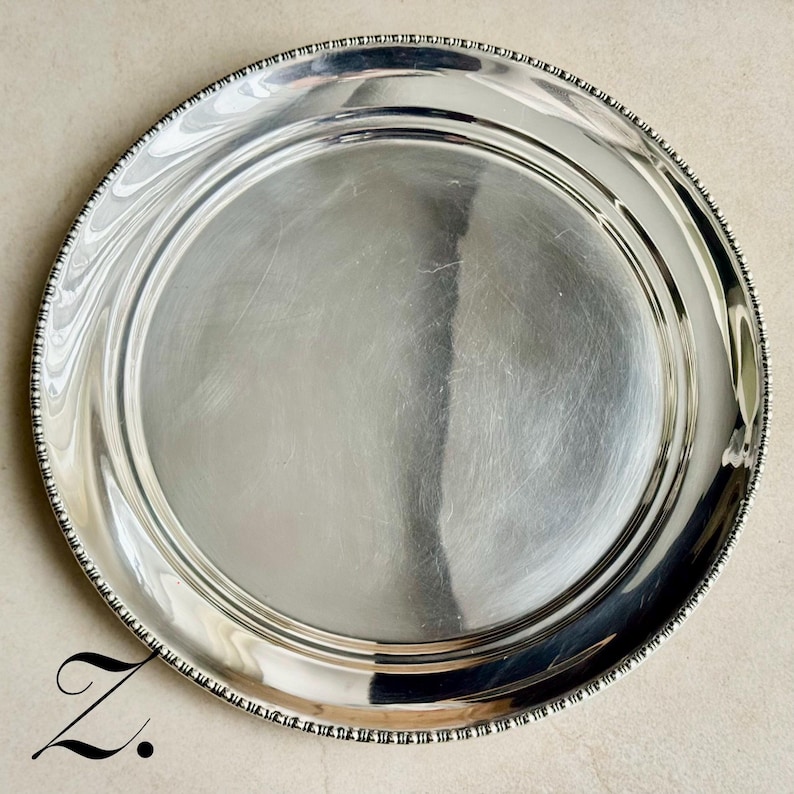 May include: A circular silver-toned metal serving tray with a polished, reflective surface and a decorative beaded edge detail. The tray features a smooth, flat center area surrounded by a slightly raised rim with a textured, repeating bead pattern.