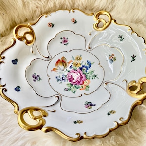 Puede incluir: Fuente de porcelana blanca con borde festoneado y ribete dorado. El plato presenta un arreglo floral central con rosas rosas y peque&ntilde;os acentos florales. El plato tiene una forma &uacute;nica con asas integradas.