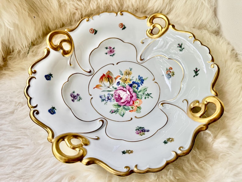 Puede incluir: Fuente de porcelana blanca con borde festoneado y ribete dorado. El plato presenta un arreglo floral central con rosas rosas y peque&ntilde;os acentos florales. El plato tiene una forma &uacute;nica con asas integradas.