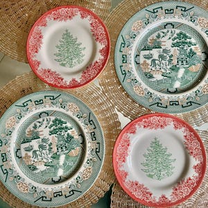 May include: Four decorative plates with intricate designs. Two plates feature a Christmas tree design with red floral borders. The other two plates have a green and white landscape scene with a decorative border. The plates are on a gold placemat.