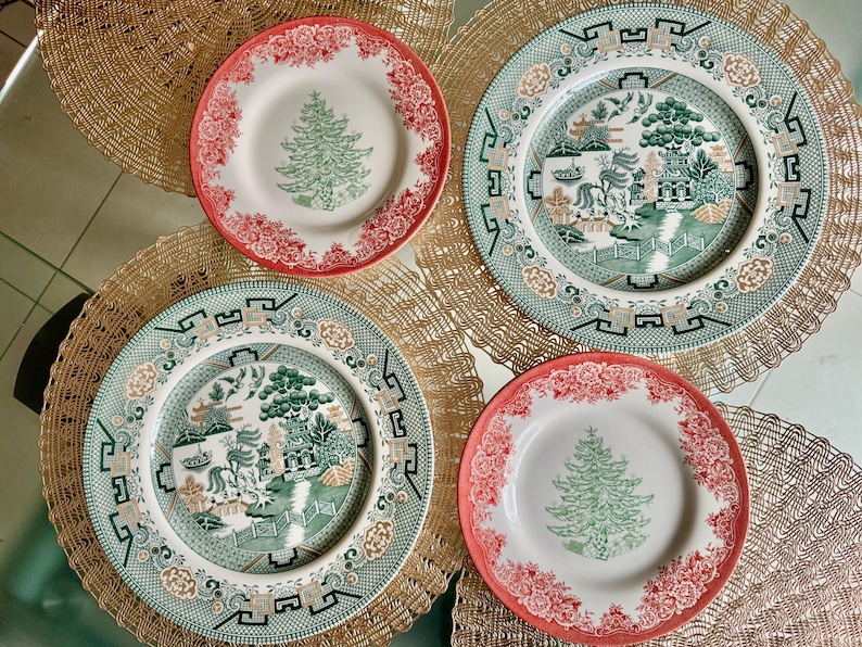 May include: Four decorative plates with intricate designs. Two plates feature a Christmas tree design with red floral borders. The other two plates have a green and white landscape scene with a decorative border. The plates are on a gold placemat.