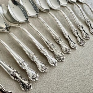 May include: A collection of silver spoons arranged diagonally. The spoons have a reflective, polished surface and ornate floral detailing on the handles. The spoons are set against a neutral background, highlighting their elegant design.