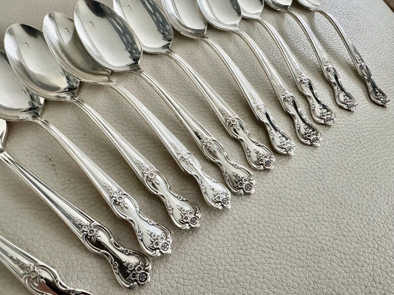 May include: A collection of silver spoons arranged diagonally. The spoons have a reflective, polished surface and ornate floral detailing on the handles. The spoons are set against a neutral background, highlighting their elegant design.