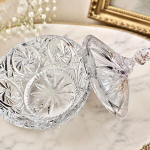 May include: A clear, cut-glass candy dish with a matching lid. The bowl and lid feature intricate, geometric starburst patterns and faceted edges that catch the light. The lid includes a pointed, decorative finial handle at the top.