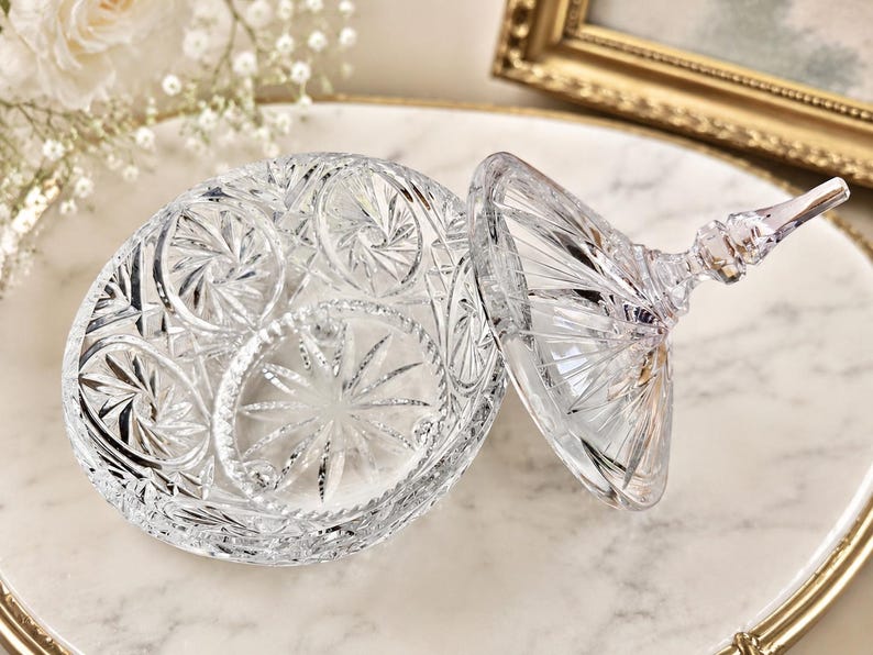May include: A clear, cut-glass candy dish with a matching lid. The bowl and lid feature intricate, geometric starburst patterns and faceted edges that catch the light. The lid includes a pointed, decorative finial handle at the top.