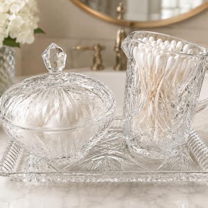 May include: A clear glass vanity set featuring a rectangular tray with a textured rim, a lidded round jar with a faceted finial, and a small pitcher filled with white cotton swabs. The glass pieces have a detailed, pressed geometric pattern.