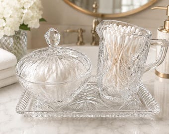 May include: A clear glass vanity set featuring a rectangular tray with a textured rim, a lidded round jar with a faceted finial, and a small pitcher filled with white cotton swabs. The glass pieces have a detailed, pressed geometric pattern.