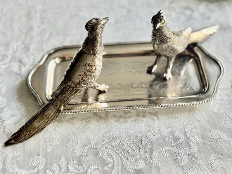 May include: A silver-toned serving tray with two bird-shaped salt and pepper shakers. The birds are detailed with feathers and stand on the tray. The tray has a decorative edge and is placed on a white patterned surface.
