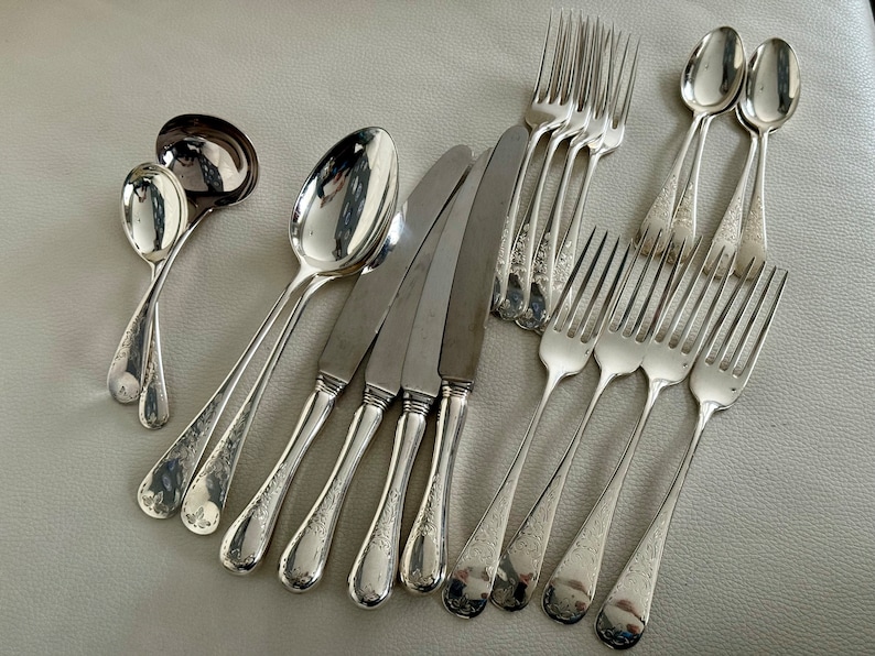 May include: A collection of antique silverware, including spoons, forks, knives, and a ladle. The silverware has a shiny, reflective surface and a floral pattern on the handles. The arrangement suggests a formal dining setting.