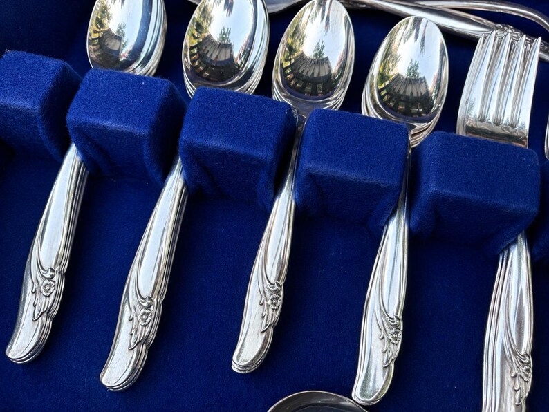 May include: A set of polished silver-toned flatware featuring spoons and a fork with embossed floral detailing on the handles. The utensils are arranged in a storage case lined with deep blue velvet, highlighting the reflective metallic surfaces.
