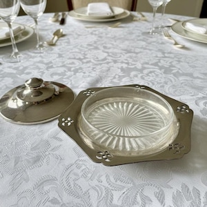 May include: A silver-toned butter dish with a clear glass insert and lid, set on a white tablecloth. The dish has a decorative, octagonal base with floral cutouts. The lid is round with a small handle.