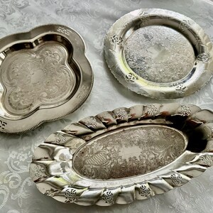 May include: Three antique silver serving trays with ornate designs. The trays vary in shape: one is clover-shaped, another is round, and the third is oval. Each tray features detailed embossed patterns and a reflective surface, showcasing a vintage aesthetic.
