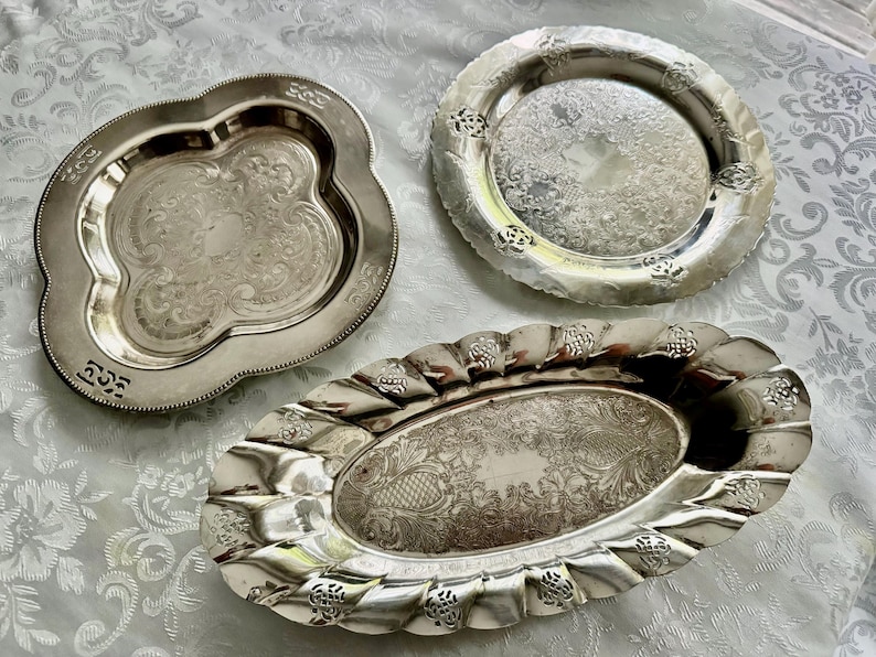 May include: Three antique silver serving trays with ornate designs. The trays vary in shape: one is clover-shaped, another is round, and the third is oval. Each tray features detailed embossed patterns and a reflective surface, showcasing a vintage aesthetic.