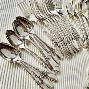 Op de afbeelding: Een assortiment antiek zilver bestek, waaronder lepels en vorken, gerangschikt op een witte, getextureerde ondergrond. Het bestek heeft sierlijke bloemendetails op de handvatten en een gepolijste afwerking. Een verzameling vintage eetgerei.