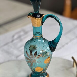 May include: A teal blue glass bottle with a gold rim and a gold floral design. The bottle has a handle and a stopper.
