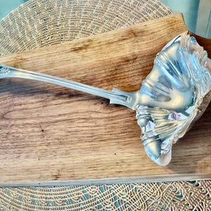 May include: A silver serving ladle with an ornate, scalloped design. The ladle is resting on a wooden surface.