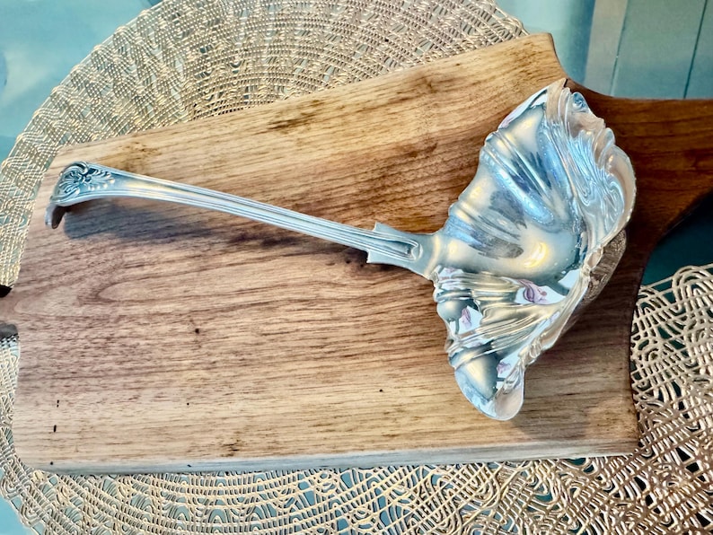 May include: A silver serving ladle with an ornate, scalloped design. The ladle is resting on a wooden surface.