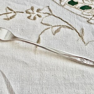 Op de afbeelding: Een zilveren vork met drie tanden, liggend op een wit linnen tafelkleed met geborduurde bloemmotieven. De vork heeft een bloemmotief aan het uiteinde. De algehele uitstraling is vintage en elegant.