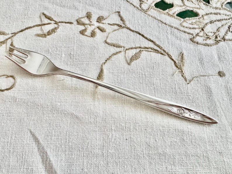 Op de afbeelding: Een zilveren vork met drie tanden, liggend op een wit linnen tafelkleed met geborduurde bloemmotieven. De vork heeft een bloemmotief aan het uiteinde. De algehele uitstraling is vintage en elegant.