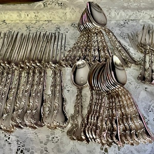 May include: A collection of ornate silver-colored flatware, including forks and spoons, arranged on a white lace tablecloth. The silverware has detailed, decorative handles. The spoons have a circular emblem on the bowl.