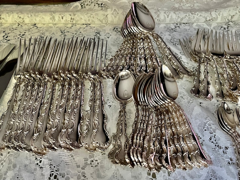 May include: A collection of ornate silver-colored flatware, including forks and spoons, arranged on a white lace tablecloth. The silverware has detailed, decorative handles. The spoons have a circular emblem on the bowl.
