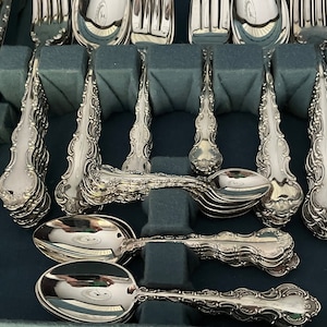 May include: A collection of ornate silver-colored flatware, including forks, spoons, and knives, arranged in a dark blue storage case. The silverware features detailed, decorative handles and is neatly organized for display or storage.
