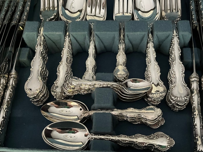 May include: A collection of ornate silver-colored flatware, including forks, spoons, and knives, arranged in a dark blue storage case. The silverware features detailed, decorative handles and is neatly organized for display or storage.
