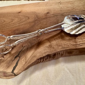 May include: Silver serving tongs with an ornate design. The tongs have a long handle and a flat, oval-shaped head with a scalloped edge.