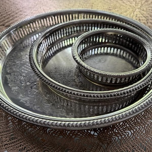 May include: A set of three nested round silver-tone metal trays with openwork gallery rails and rope-style edges. Each tray features a polished, reflective surface with an intricate, embossed floral pattern in the center.