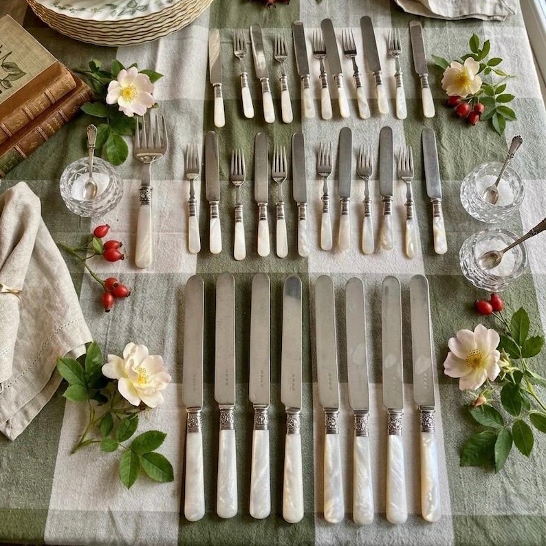 May include: An assortment of vintage silverware, including forks and knives, arranged on a green and white checkered tablecloth. The silverware features white handles and ornate silver detailing. The scene is decorated with flowers and small glass bowls.