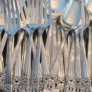 May include: A collection of polished silver-toned metal forks and spoons arranged in a cluster. Each piece features a long handle with vertical fluted lines and a decorative floral motif at the base, ending in a flat, squared-off edge.