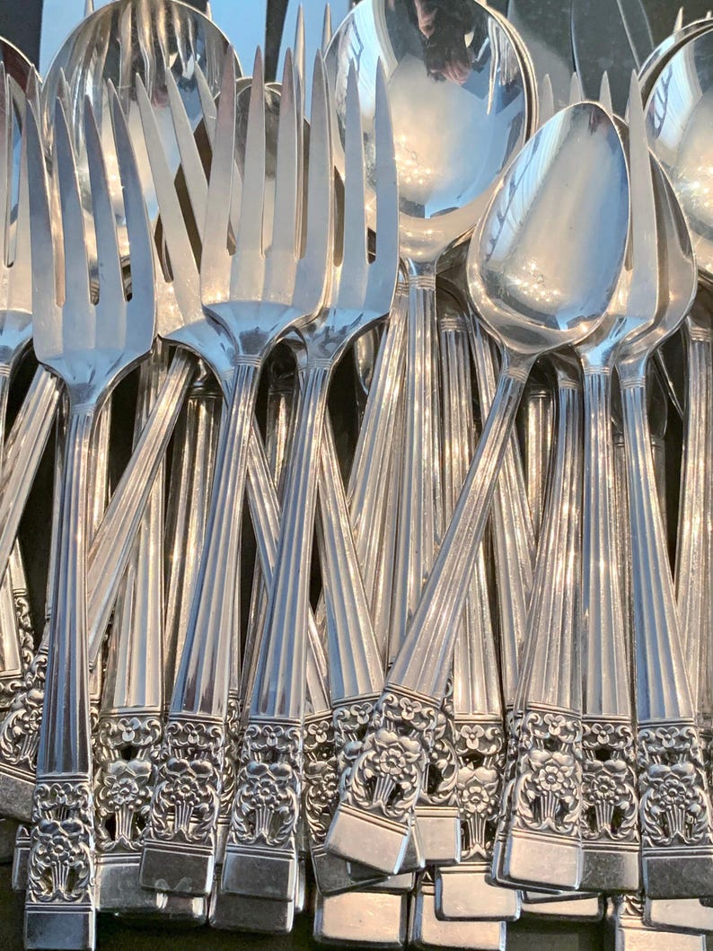 May include: A collection of polished silver-toned metal forks and spoons arranged in a cluster. Each piece features a long handle with vertical fluted lines and a decorative floral motif at the base, ending in a flat, squared-off edge.