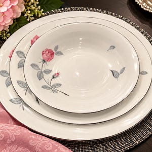 May include: A set of white ceramic dinnerware consisting of a large dinner plate, a smaller salad plate, and a bowl. Each piece features a delicate pattern of pink rose blossoms with grey leaves and a thin silver rim around the edges.