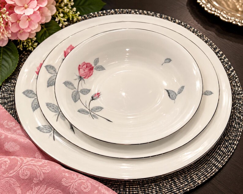 May include: A set of white ceramic dinnerware consisting of a large dinner plate, a smaller salad plate, and a bowl. Each piece features a delicate pattern of pink rose blossoms with grey leaves and a thin silver rim around the edges.