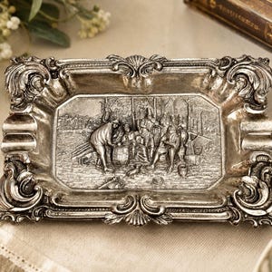 May include: A rectangular silver-toned metal ashtray featuring an ornate, raised border with scrollwork and shell motifs. The center depicts a detailed relief scene of several figures gathered around a barrel in an outdoor setting.