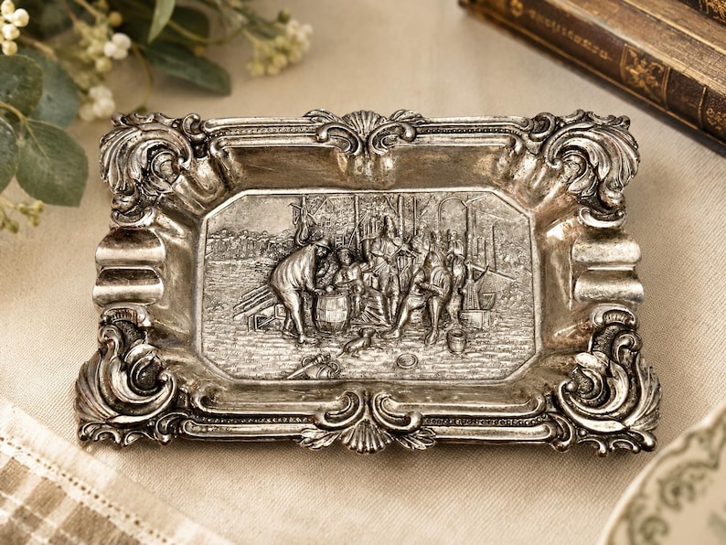 May include: A rectangular silver-toned metal ashtray featuring an ornate, raised border with scrollwork and shell motifs. The center depicts a detailed relief scene of several figures gathered around a barrel in an outdoor setting.