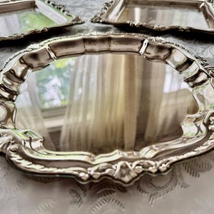 Puede incluir: Un juego de tres bandejas de servicio de metal plateado con superficies reflectantes y bordes festoneados adornados con detalles en relieve de conchas y volutas. El conjunto incluye una bandeja circular grande y dos bandejas rectangulares m&aacute;s peque&ntilde;as con bordes a juego.