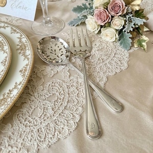 May include: Silver serving spoon and fork crossed on a lace tablecloth. The spoon has a slotted bowl. A floral arrangement of pink and white roses is in the background. Elegant table setting.